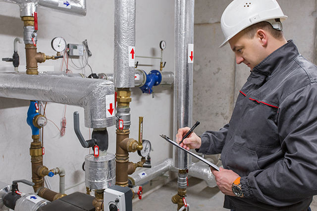 A technician with a clipboard recording readings on insulated hydronic pipework